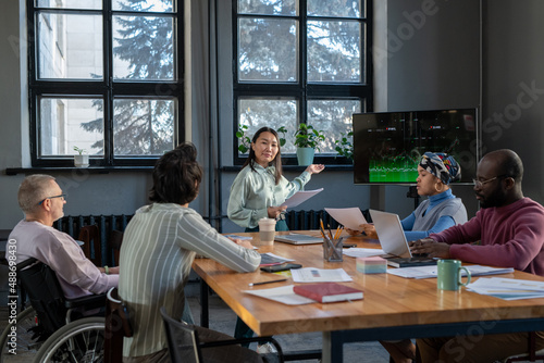 Young Asian businesswoman with documents pointing at interactive screen with graph while explaining it to group of colleagues