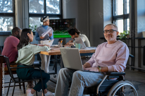 Contemporary mature business leader in wheelchair looking at camera while networking on laptop against multi-ethnic group of employees