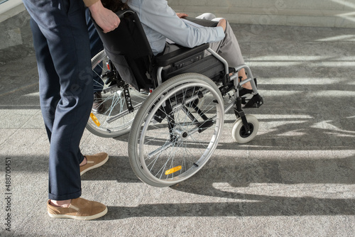 Young elegant businessman standing behind wheelchair with female colleague while helping her move inside modern office center