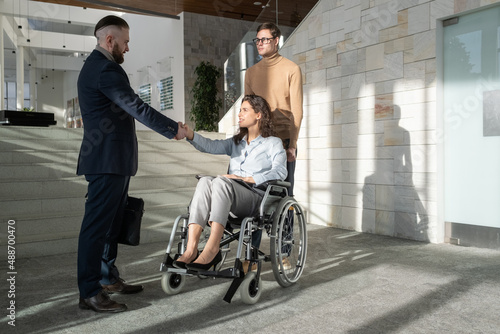 Young businesswoman with disability sitting in wheelchair while handshaking with partner after discussion of new strategies at working meeting