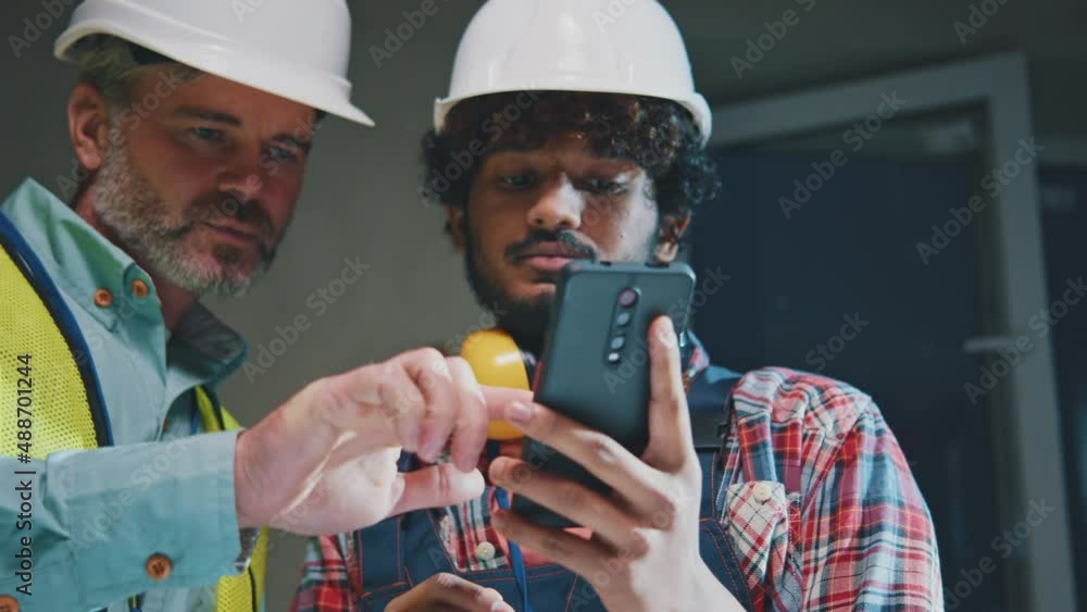 Handsome foreman and builder men in hardhats working talking using smartphone in the evening light. On table blueprints during renovation in building. Slow motion