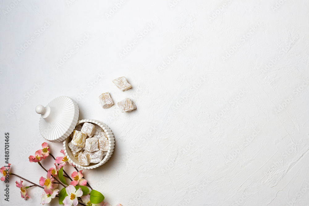 Turkish delight in a white vase with a grooved lid on a white table. A sprig of pink flowers next to a vase. Top view, space for text Turkish delight in a white vase with a grooved lid on a white table. A sprig of pink flowers next to a vase. Top view, space for text
