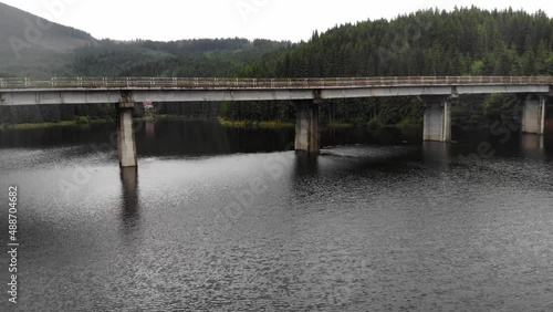drone shot flying under steel and concrete beam bridge over the Oasa Lake, Romania, part of the transalpine highway. drone goes below the bridge. Huge lake surrounded by pure green pine forest.