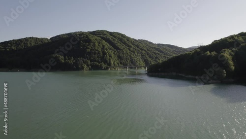 Aerial view, approaching steel and concrete beam bridge over Oasa Lake, Romania, part of the transalpine highway. drone camera aproaches the bridge. Huge lake surrounded by pure green pine forest.