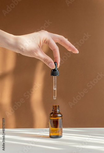 Vertical image of pipette with cosmetic serum in women's hands on light brown background, in rays of sunlight. Concept of body care, rejuvenation