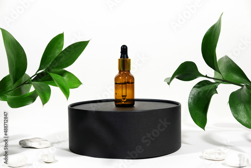 Fototapeta Naklejka Na Ścianę i Meble -  Mock up glass dropper bottle on a black podium on a white gray background with hard shadows, with leaves and stones. Hard shadows. Cosmetic pipette on a white background.