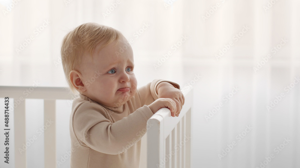 Crying baby portrait. Little infant tired and hungry, start crying standing in crib, looking aside at empty space - obrazy, fototapety, plakaty 