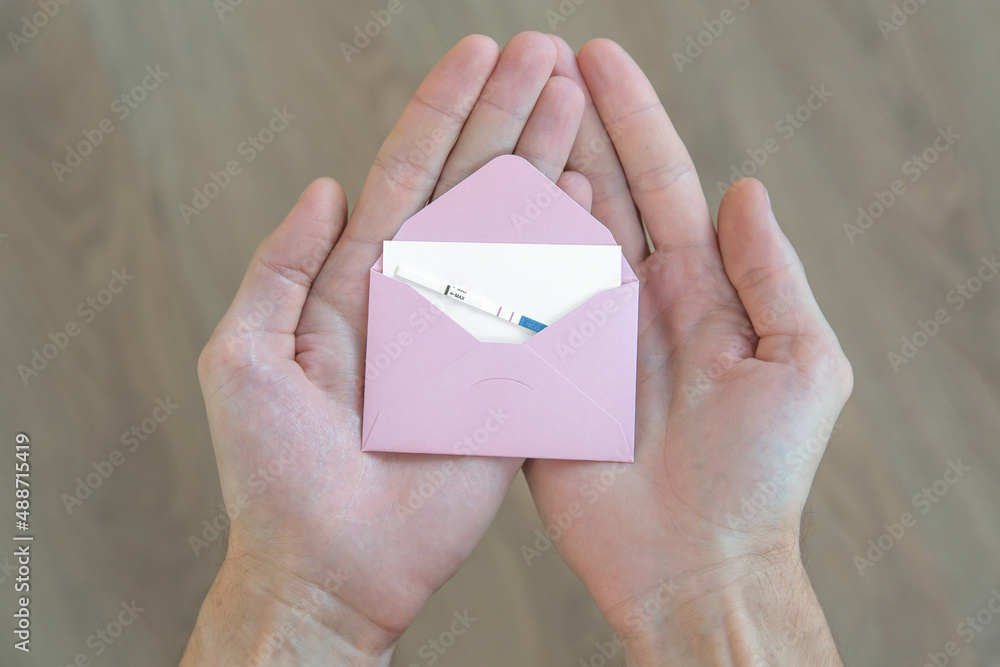 male hands hold a pink envelope with a white sheet of paper and a positive pregnancy test male hands hold a pink envelope with a white sheet of paper and a positive pregnancy test