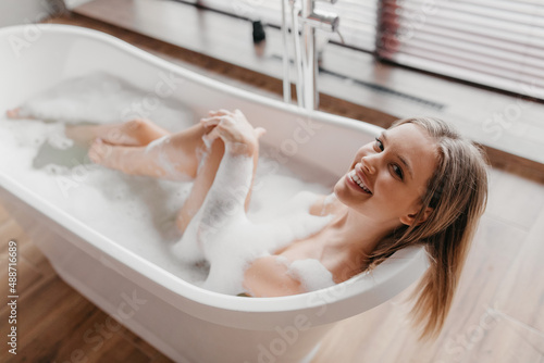 Relaxed young woman taking bath with foam  smiling at camera  resting in hot bathtub in morning at home
