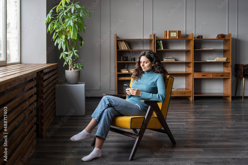 Smiling young woman rest after work in office free space or at home sit in armchair communicating with friends in smartphone app. Girl with mobile phote browsing social media or do online shopping Smiling young woman rest after work in office free space or at home sit in armchair communicating with friends in smartphone app. Girl with mobile phote browsing social media or do online shopping