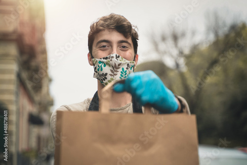 The delivery boy shows the package with the takeaway food holding it with gloves and smiling through the coronavirus protective mask photo