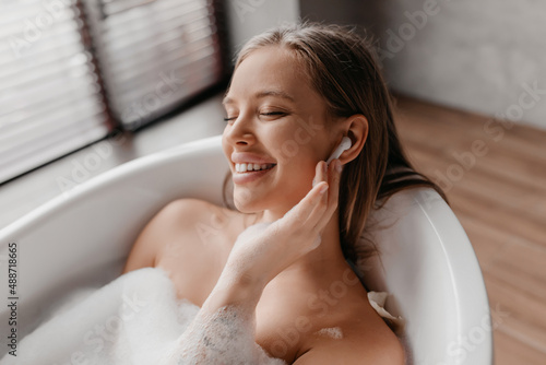 Young lady relaxing in foamy bathtub and listening music in wireless earphones, resting in bathroom at home