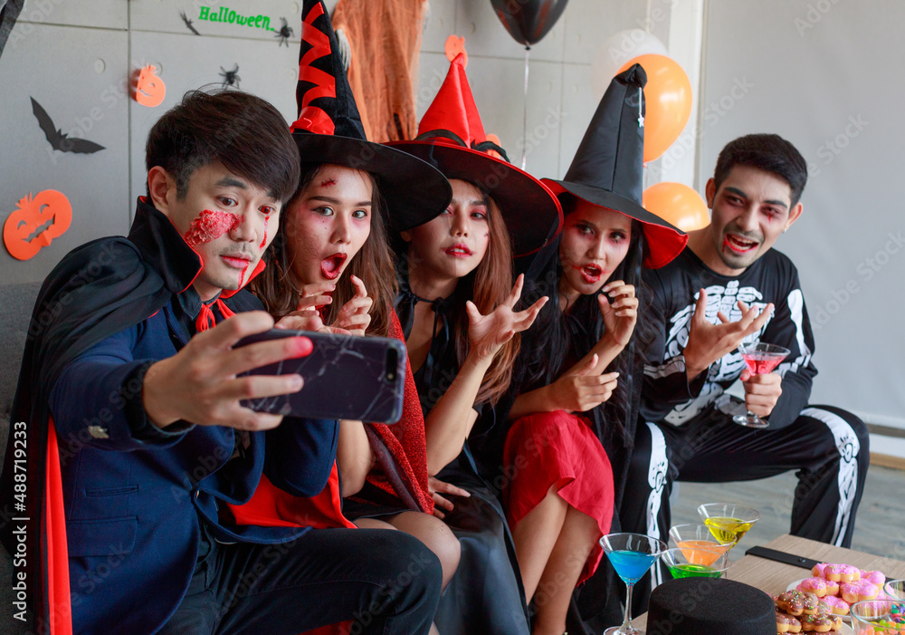 Fototapeta premium Group of Asian young scary creepy male and female friends in Halloween witch sorcerer ghost and skeleton costume sitting on sofa taking selfie photo together in traditional trick or treat night party