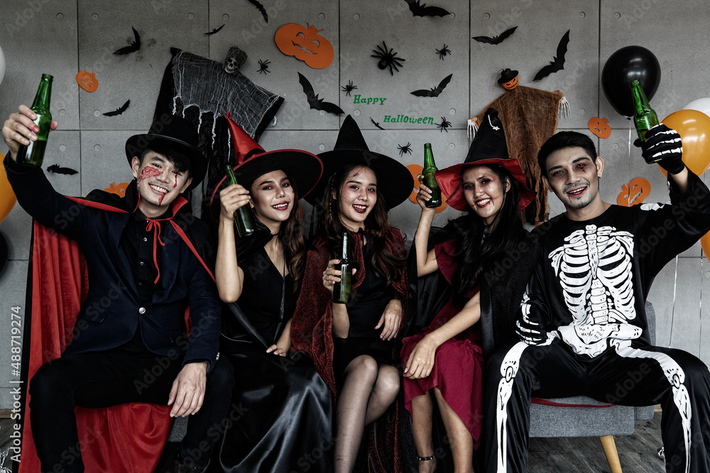 Obraz premium Portrait shot group of Asian young happy male and female friends in Halloween witch sorcerer ghost and skeleton costume with tall hat sitting on sofa holding beer bottles cheers together in party