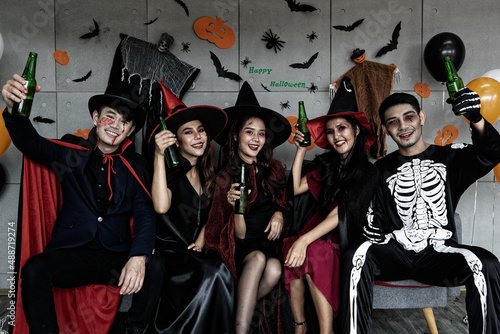 Portrait shot group of Asian young happy male and female friends in Halloween witch sorcerer ghost and skeleton costume with tall hat sitting on sofa holding beer bottles cheers together in party