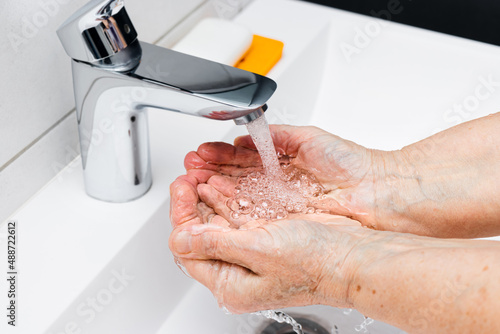 Old woman washing hands under streaming water from faucet without soap in bathroom, hygiene concept