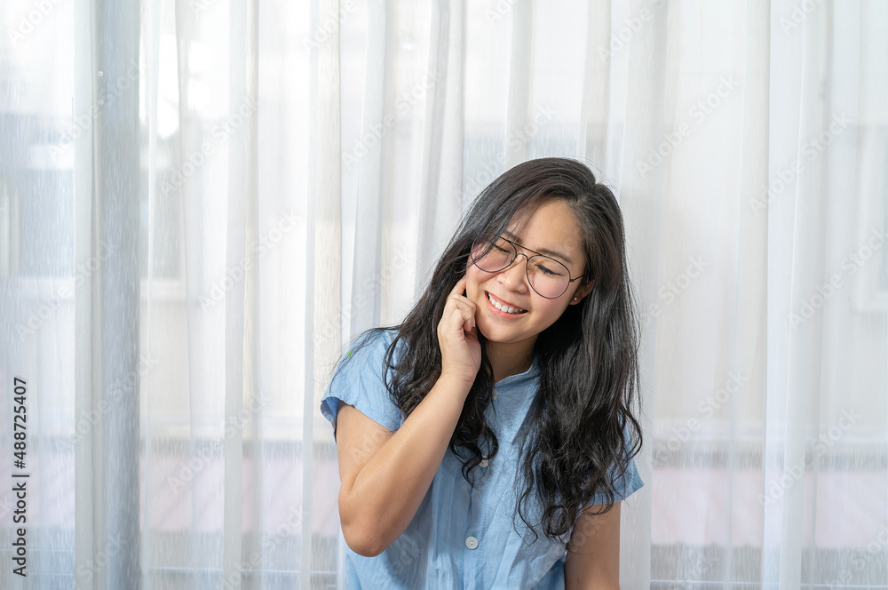 An adult Asian woman smiles as she touches her neck and cheek in front of a large window with a white curtain..