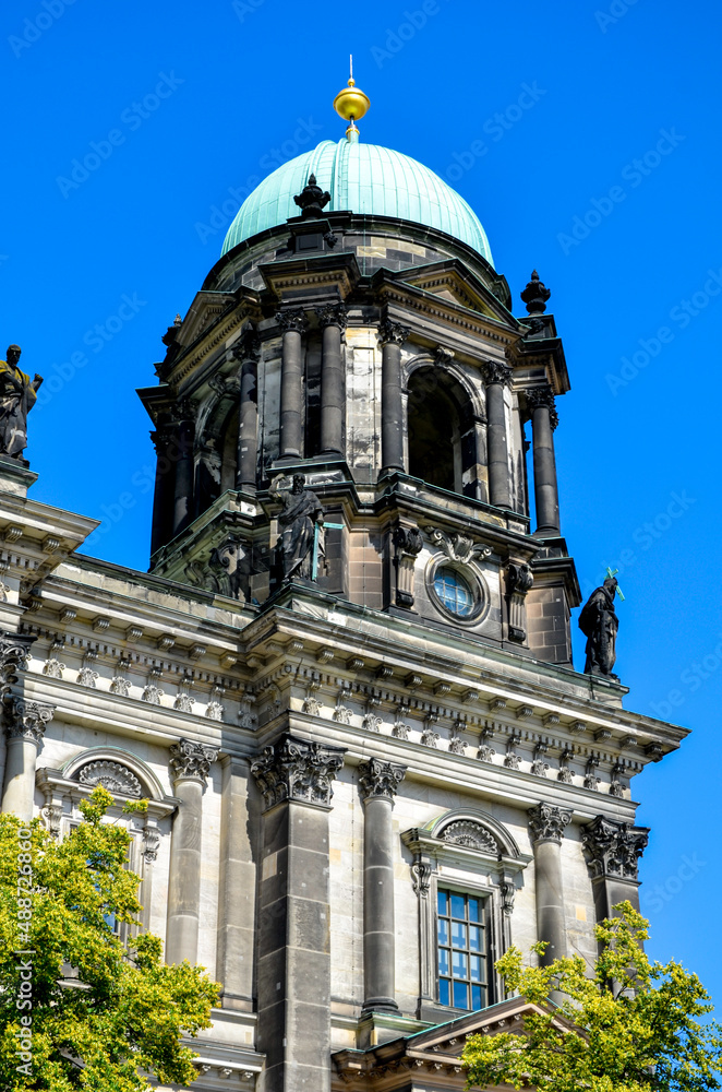 One of the outer towers of Berlin Cathedral with its green roof and some green trees in the foreground under sunny blue sky