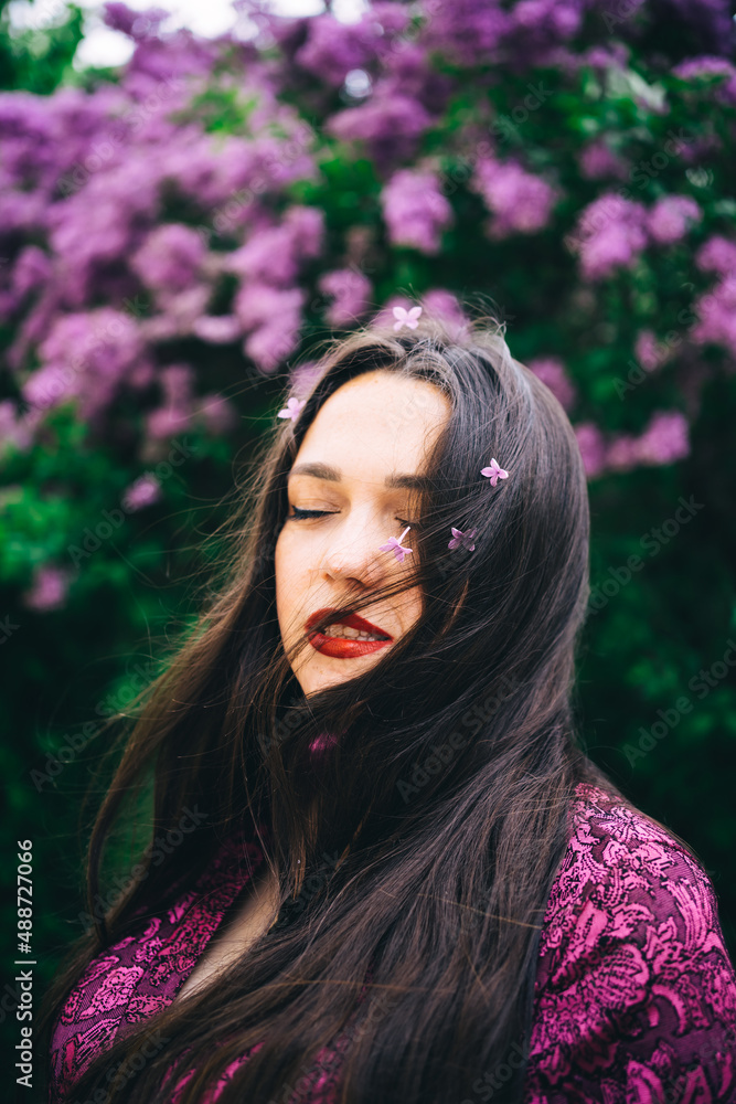 portrait of a beautiful brunette girl with flowers in her hair and red lipstick in a purple dress on a background of flowering lilac bushes