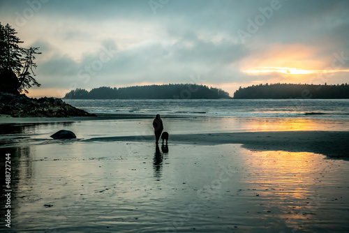 Rear view of a woman walking her dog on beach at sunset, Tofino, Vancouver Island, British Columbia, Canada photo