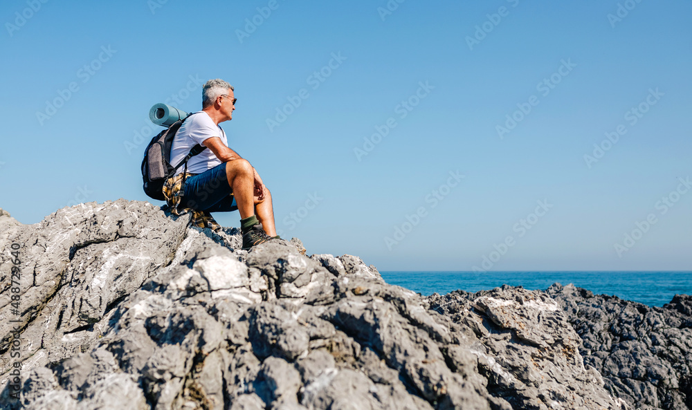 Senior man with backpack hiking looking at sea landscape sitting on rocks