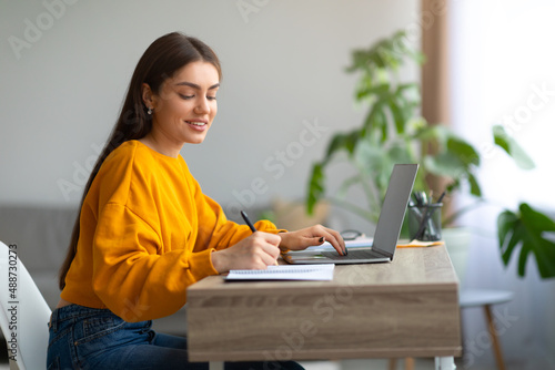 Happy Caucasian woman using laptop pc, working or studying online, taking notes during webinar or remote lesson at home