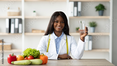 Young female african american nutritionist holding glass of water while sitting at table with fruits and vegetables © Prostock-studio