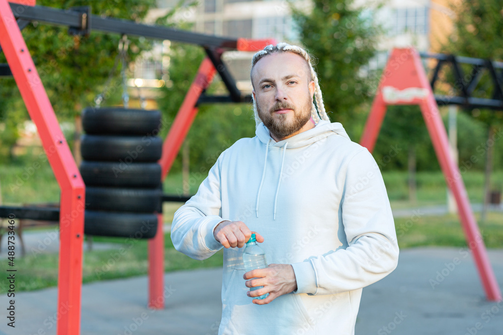 Obraz premium A man with an interesting braid hairstyle in a blue hoodie works out early in the morning on the street and drinks water from a bottle