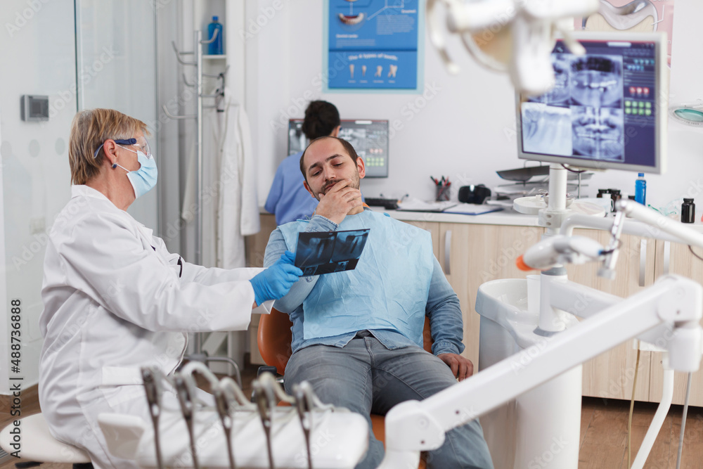 Obraz premium Senior dentist woman with face mask explaining teeth radiography to patient discussing oral treatment to prevent infection during medical examination in dental office. Stomatology concept