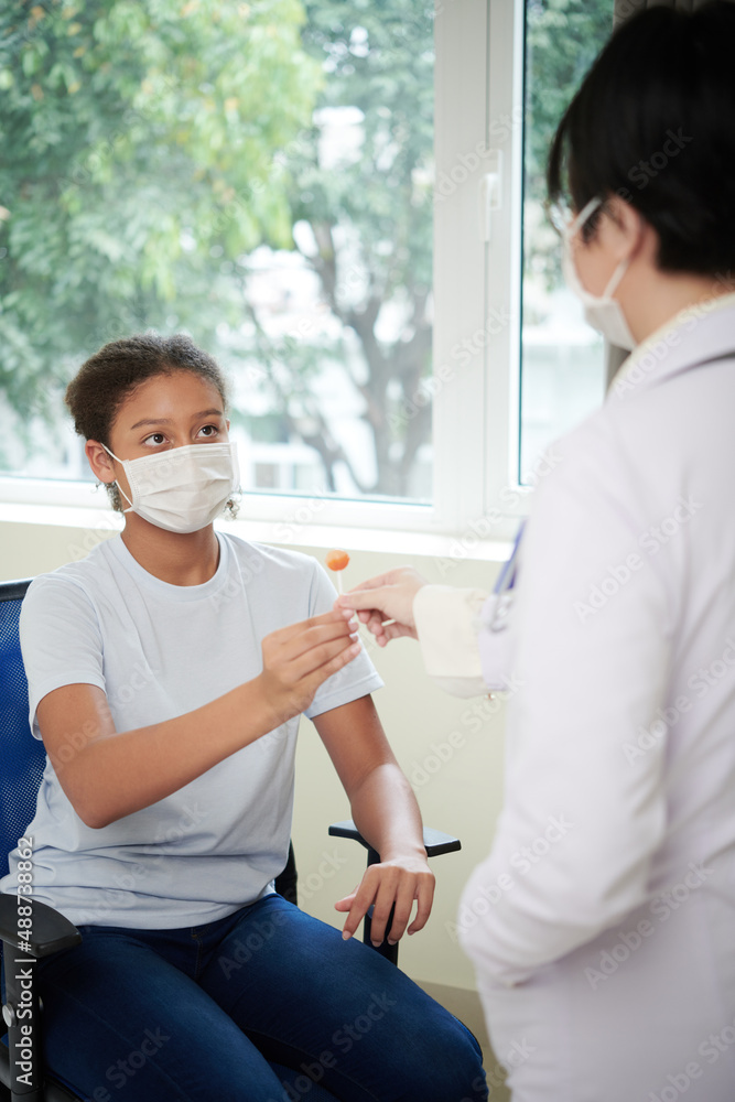 Teenage girl in mask sitting on chair and getting the candy from female doctor during medical exam at hospital