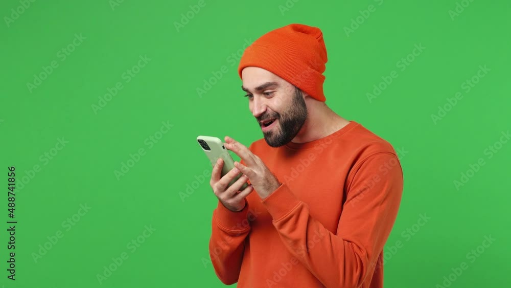 Happy fun young brunet man 20s years old wears red shirt hat hold use mobile cell phone typing browsing swipe chatting send sms doing online shopping isolated on plain light green background studio Happy fun young brunet man 20s years old wears red shirt hat hold use mobile cell phone typing browsing swipe chatting send sms doing online shopping isolated on plain light green background studio