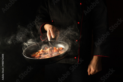 Fototapeta Naklejka Na Ścianę i Meble -  Chef prepares food in a frying pan with steam on a black background. The concept of restaurant and hotel service. European cuisine.