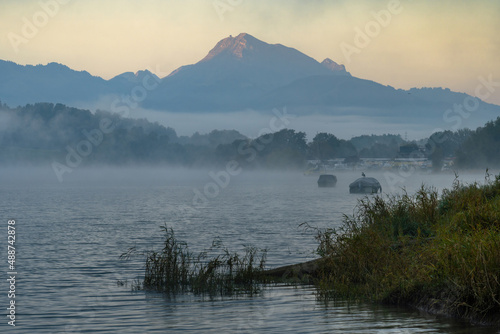 Scene photographed from the shore of a lake in the early morning of autumn. The night fog is clearing, allowing glimpses of moored boats and a few gulls. The lake water is slightly choppy and a