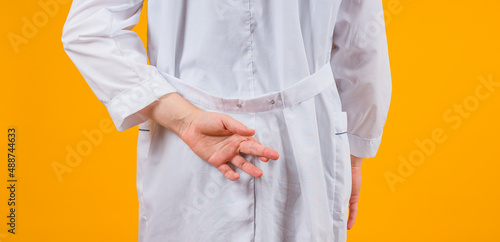 male doctor in a white coat with his fingers crossed behind his back on a yellow background, concept for good luck, close-up