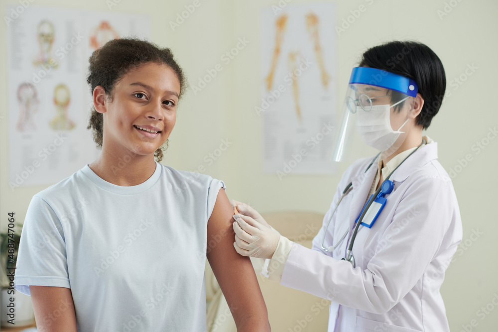 Portrait of teenage girl smiling at camera while nurse in protective wear giving a vaccination during her visit at hospital Portrait of teenage girl smiling at camera while nurse in protective wear giving a vaccination during her visit at hospital