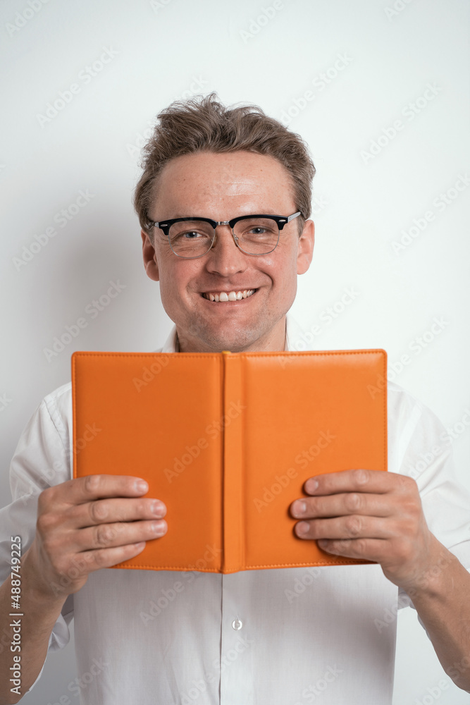 Looking out of orange notebook or journal young man in eye glasses wearing white shirt isolated on white background or in the office. Portrait of a shy young businessman holding his diary. Looking out of orange notebook or journal young man in eye glasses wearing white shirt isolated on white background or in the office. Portrait of a shy young businessman holding his diary.