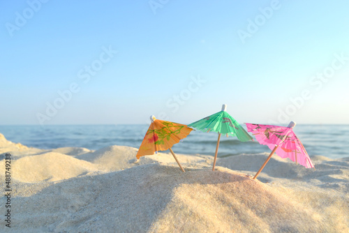 Three small paper cocktail umbrellas stand in sand on sandy beach close-up. Small paper umbrellas on sandy shore near sea and sea waves on sunny summer day. Leisure  Vacation  Travel  Tourism Concept