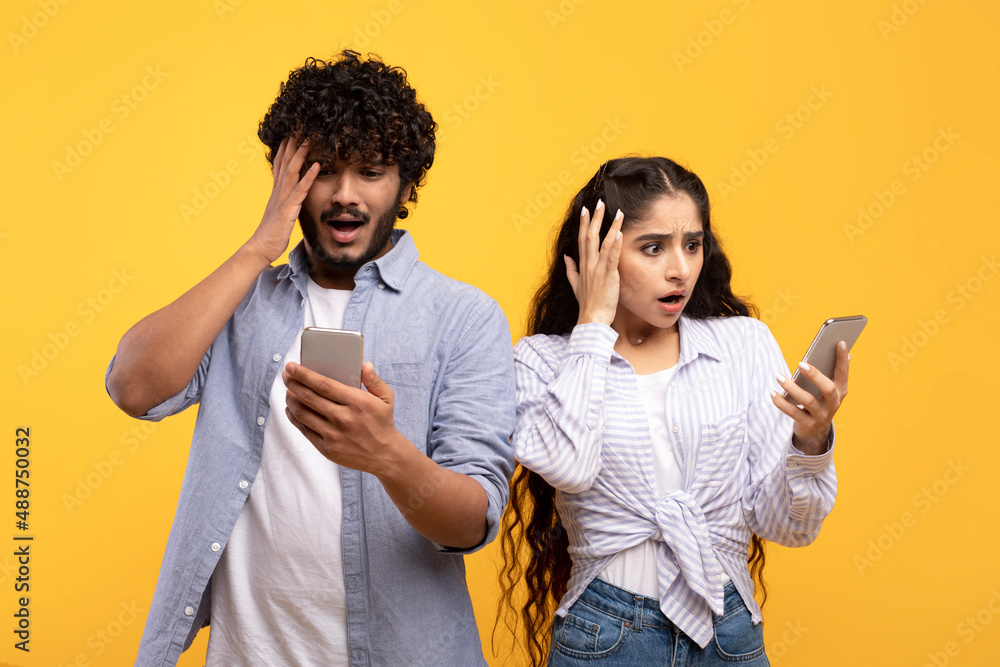 Indian woman and her boyfriend looking into their smartphones in shock and touching heads on yellow background Indian woman and her boyfriend looking into their smartphones in shock and touching heads on yellow background