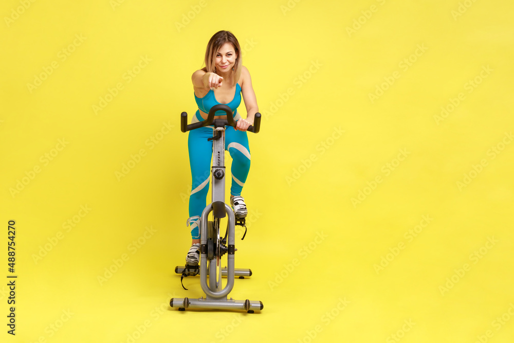 Full length portrait of blonde young adult sporty woman having cardio workout on exercise bike, pointing finger to camera, inviting you doing sport. Indoor studio shot isolated on yellow background. Full length portrait of blonde young adult sporty woman having cardio workout on exercise bike, pointing finger to camera, inviting you doing sport. Indoor studio shot isolated on yellow background.
