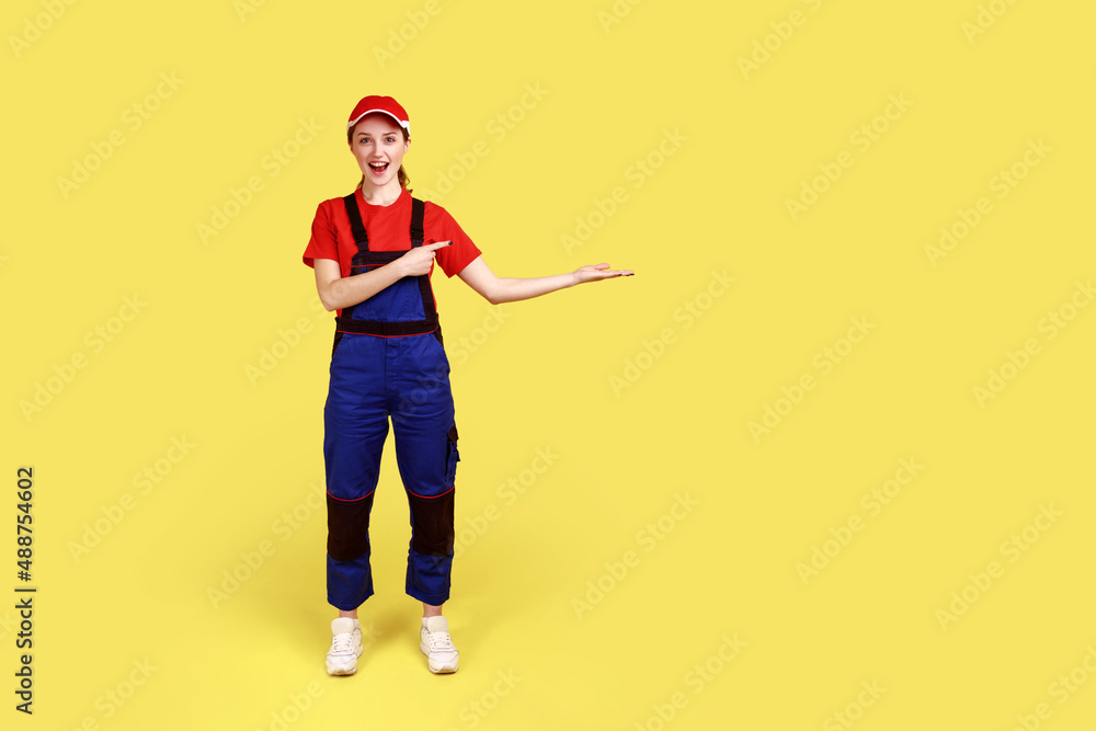 Full length portrait of amazed worker woman presenting copy space for advertisement on palm, looks at camera with excitement, wears overalls and cap. Indoor studio shot isolated on yellow background Full length portrait of amazed worker woman presenting copy space for advertisement on palm, looks at camera with excitement, wears overalls and cap. Indoor studio shot isolated on yellow background
