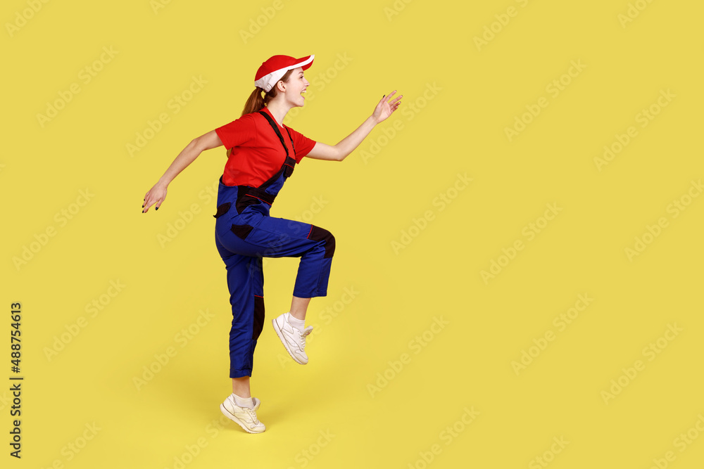 Side view of positive worker woman standing on one leg with raised arms, looking ahead, marching, happy expression, wearing overalls and red cap. Indoor studio shot isolated on yellow background Side view of positive worker woman standing on one leg with raised arms, looking ahead, marching, happy expression, wearing overalls and red cap. Indoor studio shot isolated on yellow background
