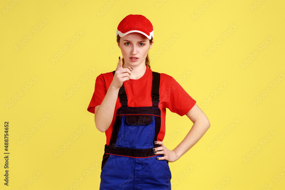 Portrait of bossy confident worker woman standing, raising finger, showing warning gesture, looking at camera, wearing overalls and red cap. Indoor studio shot isolated on yellow background. Portrait of bossy confident worker woman standing, raising finger, showing warning gesture, looking at camera, wearing overalls and red cap. Indoor studio shot isolated on yellow background.