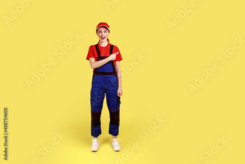 Full length portrait of excited amazed handy woman standing and pointing finger away, showing copy space for advertisement, wearing overalls and cap. Indoor studio shot isolated on yellow background.