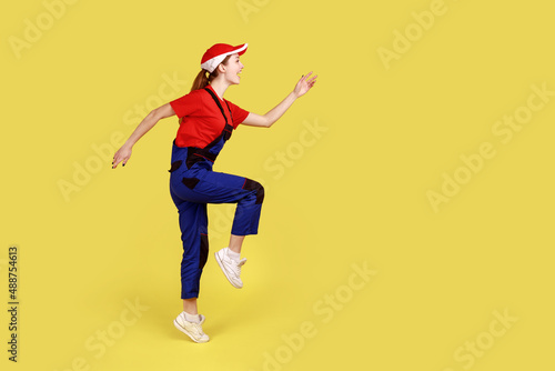 Side view of positive worker woman standing on one leg with raised arms, looking ahead, marching, happy expression, wearing overalls and red cap. Indoor studio shot isolated on yellow background