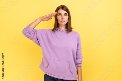 Portrait of responsible patriotic woman following discipline, saluting to commander with hand near temple and listening order, wearing purple hoodie. Indoor studio shot isolated on yellow background.