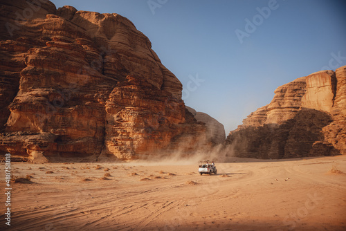 Wadi Rum desert in Jordan. A car driving in the distance on sandy desert terrain, with big rocky mountains in the distance. Wind blowing the sand and making a sand storm