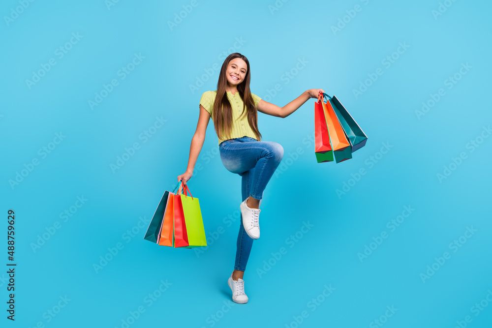 Photo of cheerful dream little lady hold store bags wear green top jeans sneakers isolated blue color background Photo of cheerful dream little lady hold store bags wear green top jeans sneakers isolated blue color background