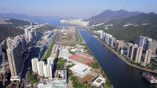 Tall ultra-modern apartments with expansive shadows next to Sha Tin race track and the Shing Mun river on a hot summer day in Hong Kong. Drone backwards dolly shot photo