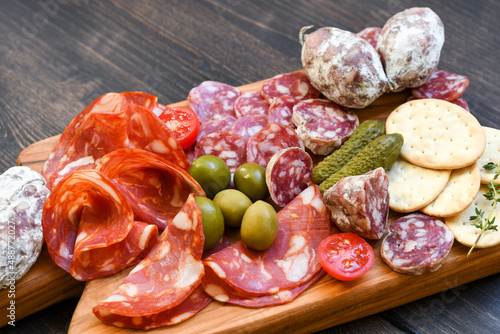 Various types of salami on a wooden board, served with olives, crackers and pickles and cracker. Food for an aperitif and dinner lunch in the restaurant. 