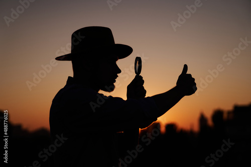 Male silhouette in hat looking with magnifying glass showing thumbs up gesture. Man searching sales or discounts at sunset using loupe with sun on background and urban view. Investigation concept